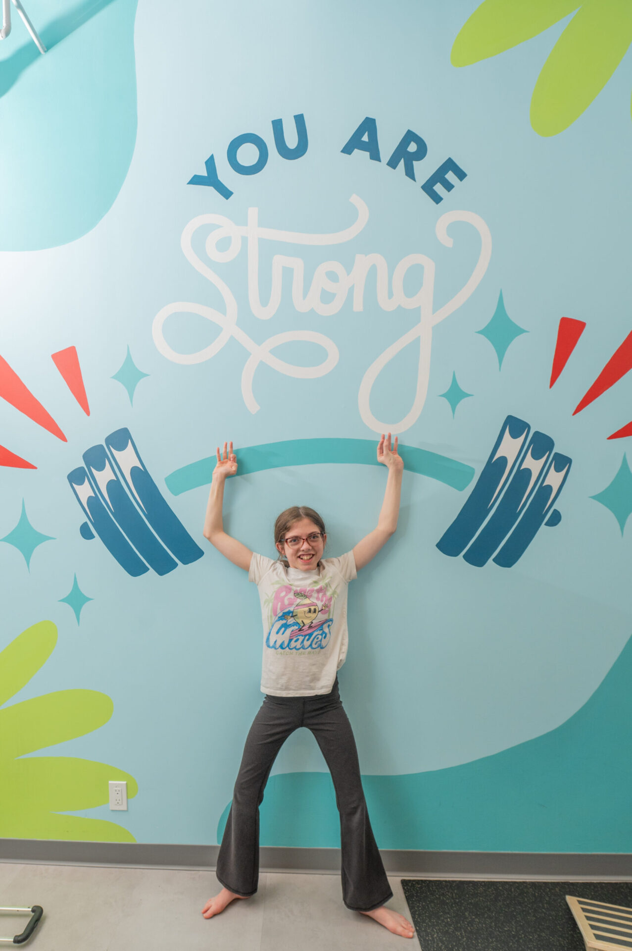 Girl poses like she’s lifting weights under mural that reads you are strong